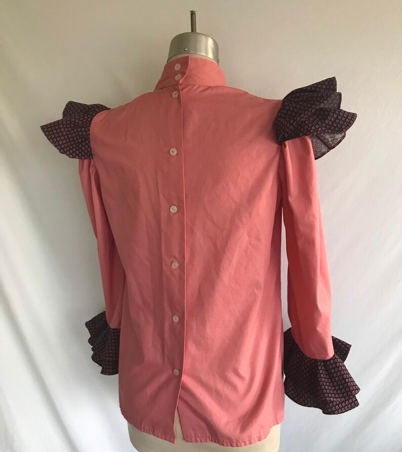 May include: A pink long-sleeved blouse with a high collar and button-up back. The sleeves and shoulders have ruffled trim in a black and white floral print.