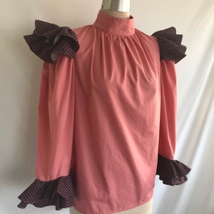 May include: A pink long-sleeve blouse with a high neck and ruffled trim on the shoulders and cuffs. The ruffles are a dark brown with a small geometric pattern.