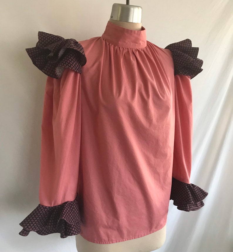 May include: A pink long-sleeve blouse with a high neck and ruffled trim on the shoulders and cuffs. The ruffles are a dark brown with a small geometric pattern.