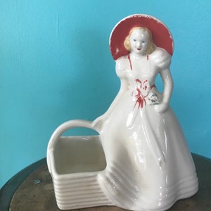 May include: White ceramic figurine of a woman wearing a red hat and holding a bouquet of flowers. The woman is sitting on a white ceramic basket.
