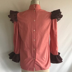 May include: A pink button-up shirt with a high collar and ruffled sleeves. The sleeves are trimmed with a black and white patterned fabric.