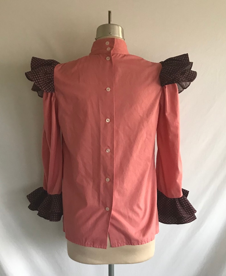May include: A pink button-up shirt with a high collar and ruffled sleeves. The sleeves are trimmed with a black and white patterned fabric.