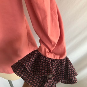 May include: A coral pink long-sleeved blouse with a double-layered ruffle at the cuffs. The ruffle is brown with a white and pink floral pattern.