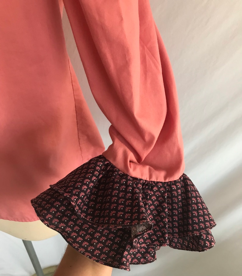 May include: A coral pink long-sleeved blouse with a double-layered ruffle at the cuffs. The ruffle is brown with a white and pink floral pattern.