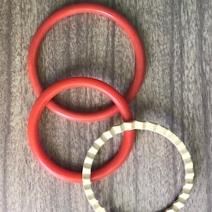 May include: Three bangles, two are orange and one is gold. The orange bangles are smooth and the gold bangle has a scalloped edge.