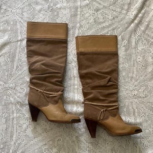 May include: A pair of brown suede knee-high boots with a stacked heel and a wraparound braided strap detail. The boots have a pointed toe and a slouchy fit.