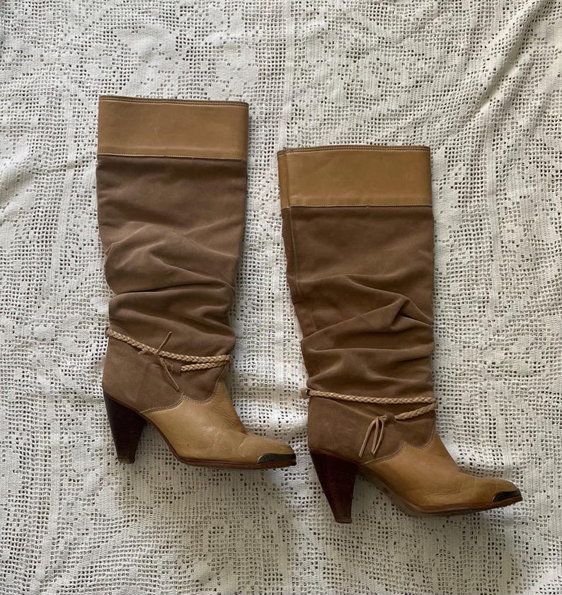 May include: A pair of brown suede knee-high boots with a stacked heel and a wraparound braided strap detail. The boots have a pointed toe and a slouchy fit.