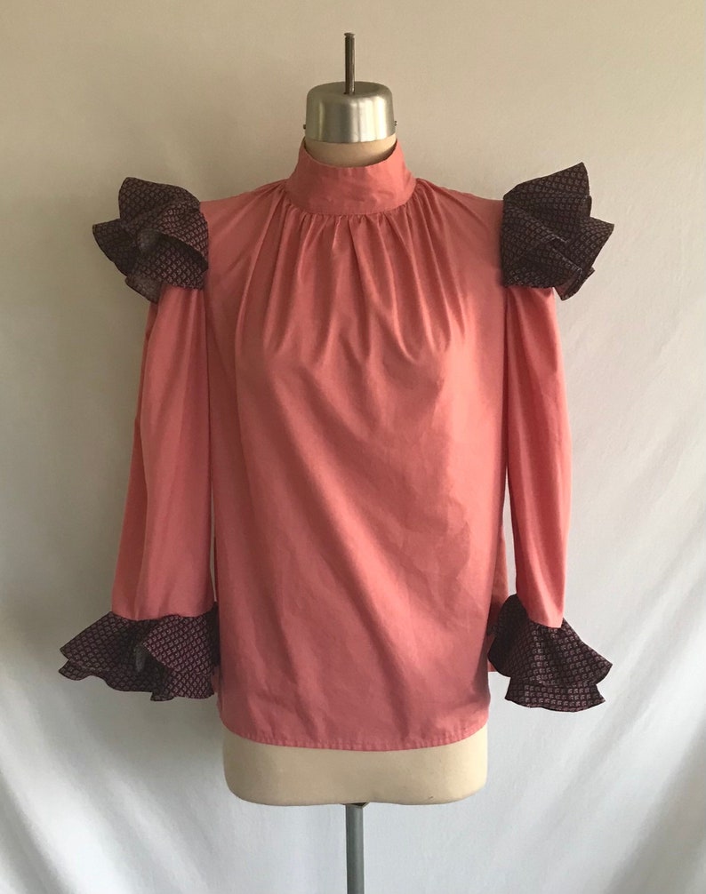 May include: A salmon pink blouse with a high neck and long sleeves. The sleeves and shoulders have black and white patterned ruffles.