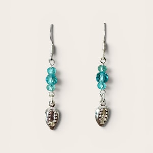 May include: A pair of dangle earrings featuring silver-toned fishhook wires, each adorned with three faceted turquoise-blue beads of varying sizes and a single metallic cowrie shell charm suspended at the bottom of the vertical arrangement.