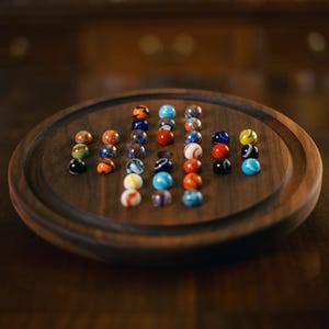 May include: A wooden board game with a circular design, featuring a collection of colorful marbles. The marbles are arranged in a pattern, showcasing various colors like blue, orange, red, and white. The board is made of dark wood.