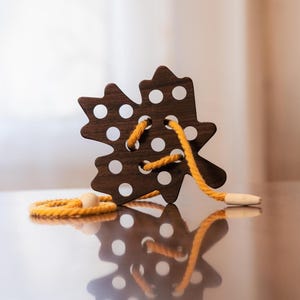 Könnte beinhalten: Ein braunes Holzspielzeug in Blattform mit weißen Punkten und einer gelben Schnur zum Schnüren. Das Spielzeug hat eine kleine Holzperle am Ende der Schnur. Das Spielzeug ist für Kinder konzipiert.
