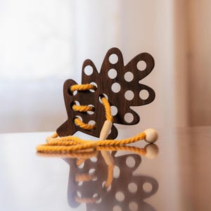 May include: A wooden leaf-shaped toy with a dark brown finish and a series of white circular cutouts. Bright yellow rope is laced through the toy, with a wooden bead at the end. The toy is designed for children.