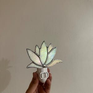 Könnte beinhalten: Ein Nachtlicht aus Buntglas in Form einer Lotusblume. Die Blume hat irisierende Blütenblätter mit silbernen Lötlinien. Der Sockel ist weiß und wird an eine Standardsteckdose angeschlossen.