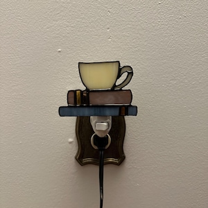 Może przedstawiać: Witrożowa lampka nocna przedstawiająca filiżankę i dwie książki. Filiżanka jest jasnożółta z ciemnym konturem, spoczywająca na dwóch książkach z brązowymi i niebieskimi okładkami. Lampka nocna jest podłączona do gniazdka ściennego.
