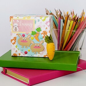 May include: A decorative box with a bunny theme, featuring the text "You are such a Funny Bunny." The box is decorated with cartoon bunnies, carrots, and colorful accents. A felt carrot and a glass of colored pencils are also visible.