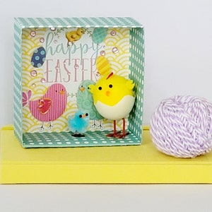 May include: A decorative Easter-themed box with a yellow base, featuring a cheerful scene of cartoon chicks and the words "Happy Easter." The box has a mint green and white polka dot pattern and a light purple ball of twine.