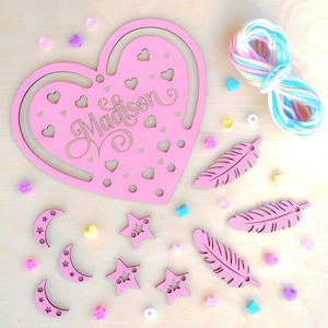 May include: Pink wooden heart-shaped craft kit with the name Madison cut out. The kit includes pink wooden stars, moons, and feathers, as well as a spool of rainbow yarn.