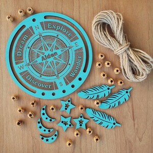 May include: A teal blue wooden compass with the words "Dream", "Explore", "Wander", and "Discover" around the compass rose. The compass also has the name "Aiden" in the center. The compass is surrounded by wooden beads, feathers, stars, and crescent moons.