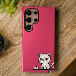 May include: A magenta phone case featuring a cartoon illustration of an angry white cat with red eyes. The case has a multi-lens camera cutout at the top and a small cup next to the cat. The case is on a wooden surface.