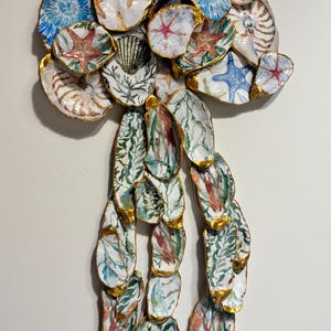 May include: A decorative wall hanging made of painted oyster shells. The shells are adorned with sea life designs, including starfish and seashells, and have gold-painted edges. The overall shape resembles a jellyfish.
