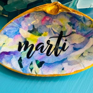 May include: A decorative seashell dish with a gold-painted rim. The interior features a watercolour floral design in shades of blue, purple, and yellow. The name "Marti" is written in black script across the centre.
