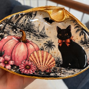 May include: A decorative seashell dish with a gold-painted rim featuring a glossy illustration of a black cat with an orange bow, a large pink pumpkin, pink flowers, a smaller seashell, a bat silhouette, and a coastal scene with palm trees.