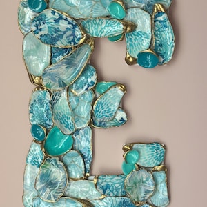 May include: A decorative letter E wall hanging constructed from numerous individual oyster shells. Each shell is painted in various shades of blue, teal, and white with intricate patterns and finished with metallic gold edges on the rims.