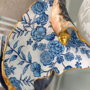 May include: A decorative seashell with a blue floral pattern on a white background. The edges are trimmed with gold paint. The shell has a natural, textured appearance with a gold-painted tip.