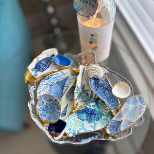 May include: A decorative glass dish with a gold rim, filled with blue and white seashell-shaped pieces. A clear glass bottle with fairy lights and a tag that says "happen, it's ok to w" is in the background.