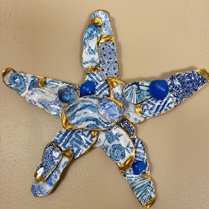 May include: A starfish-shaped wall art piece crafted from repurposed blue and white porcelain shards. The edges are outlined in gold, and blue seashell accents are interspersed throughout. The porcelain features floral and geometric patterns, creating a unique coastal decor item.