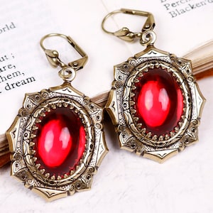 May include: A pair of antique-style earrings with a bronze finish. Each earring features a large, oval, ruby-red gemstone set within an ornate, filigree frame. The earrings have lever-back closures.