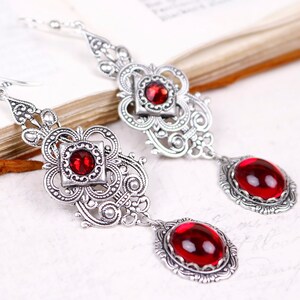 May include: Silver-toned dangle earrings with ornate filigree detailing. Each earring features a square red gemstone at the top and a large oval red gemstone at the bottom. The earrings are displayed on an open book.