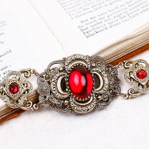 May include: An antique-style bracelet with a bronze finish and vibrant red gemstones. The bracelet features an ornate, filigree design with a central oval stone and smaller round and oval stones. The clasp is a lobster claw style.