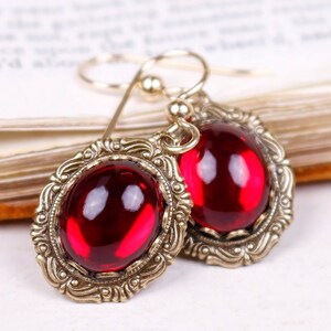 May include: A pair of vintage-style earrings featuring large, oval, ruby-red gemstones set in ornate, antique gold-toned metal frames. The earrings have a hook closure and are displayed against a blurred background.