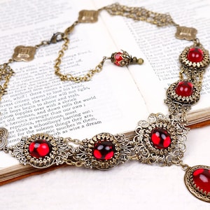 May include: An antique-style necklace with a bronze-colored chain and ornate filigree settings. The necklace features large, oval, and round red glass cabochons. The design includes a pendant and multiple strands, creating a vintage aesthetic.