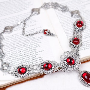 May include: A silver-toned necklace with ornate filigree and vibrant red gemstone accents. The necklace features a series of round and oval red stones, set within detailed metalwork, and is displayed on an open book.