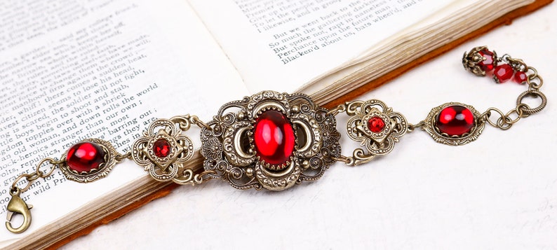 Pu&ograve; includere: Un bracciale in stile antico con finitura bronzo e pietre preziose rosse vibranti. Il bracciale presenta un design a filigrana ornato con una pietra ovale centrale e pietre rotonde e ovali pi&ugrave; piccole. La chiusura &egrave; a moschettone.