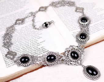 May include: Silver-toned necklace with black oval and round cabochons. The necklace features an ornate, filigree design with a central pendant and a delicate chain. The necklace is displayed on an open book.