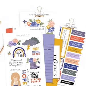 Op de afbeelding: Een verzameling papieren ephemera met bloemen- en inspirerende ontwerpen. De ontwerpen omvatten een regenboog, bloemen en tekst zoals "Hope Anchors the Soul" en bijbelverzen. De papieren worden bij elkaar gehouden met gouden paperclips.