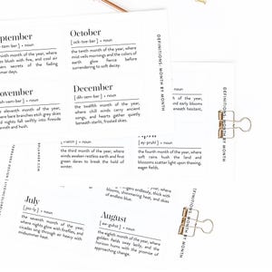 Könnte beinhalten: Weiße Karten mit schwarzem Text, die jeweils einen Monat des Jahres und seine Definition zeigen. Goldene Büroklammern halten die Karten zusammen. Die Karten stammen von einem Planer und haben ein minimalistisches Design.