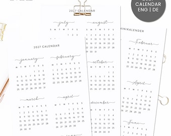 May include: A 2027 wall calendar printed on white paper with black text, featuring months from January to December in English. The calendar is held together by a gold metal binder clip at the top, displaying a clean, minimalist layout.