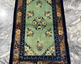 Op de afbeelding: Een vintage Chinees tapijt met een groen midden en een blauwe, oranje en witte rand. Het tapijt heeft een bloemmotief met ingewikkelde details.