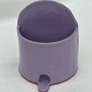 Peut inclure: Un petit porte-brosse à dents en céramique de couleur lavande avec un dessus arrondi ouvert et un seul pied arrondi.
