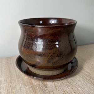 May include: A brown ceramic planter with a matching saucer. The planter has a speckled glaze and a rounded shape.