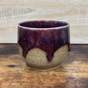 May include: A handmade ceramic cup with a speckled tan base and a deep burgundy glaze that drips down the sides. The rim has a lighter, almost white, color. The cup sits on a wooden surface.