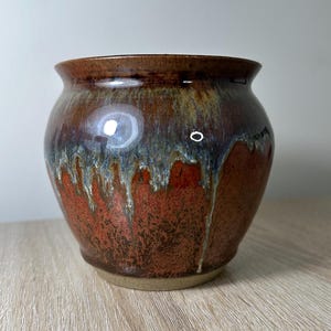 May include: A brown ceramic pot with a drip glaze in shades of red, brown, and blue. The pot has a rounded shape and a slightly flared rim.