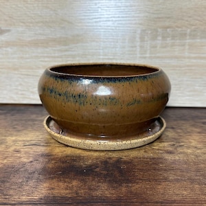 May include: A brown ceramic planter with a matching saucer. The planter has a rounded shape with a dark blue rim and a speckled, textured surface. The saucer is a lighter brown with a speckled pattern.