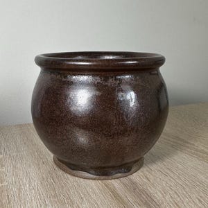 May include: A dark brown ceramic pot with a rounded body and a slightly flared rim. The pot has a speckled glaze and a slightly uneven finish.