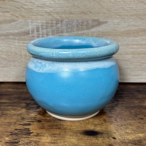 May include: A small, round ceramic pot with a light blue exterior and a slightly darker blue rim. The pot has a white band near the top and sits on a white base. The surface has a crackle glaze.