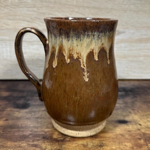 May include: A handmade ceramic mug with a brown glaze that drips down from the rim. The mug has a rounded body, a sturdy handle, and a light-colored base. The mug is designed for hot beverages.